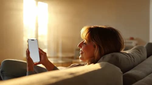 Frau stöbert entspannt lächelnd auf dem Sofa per Smartphone durch Video-Shopping-Angebote